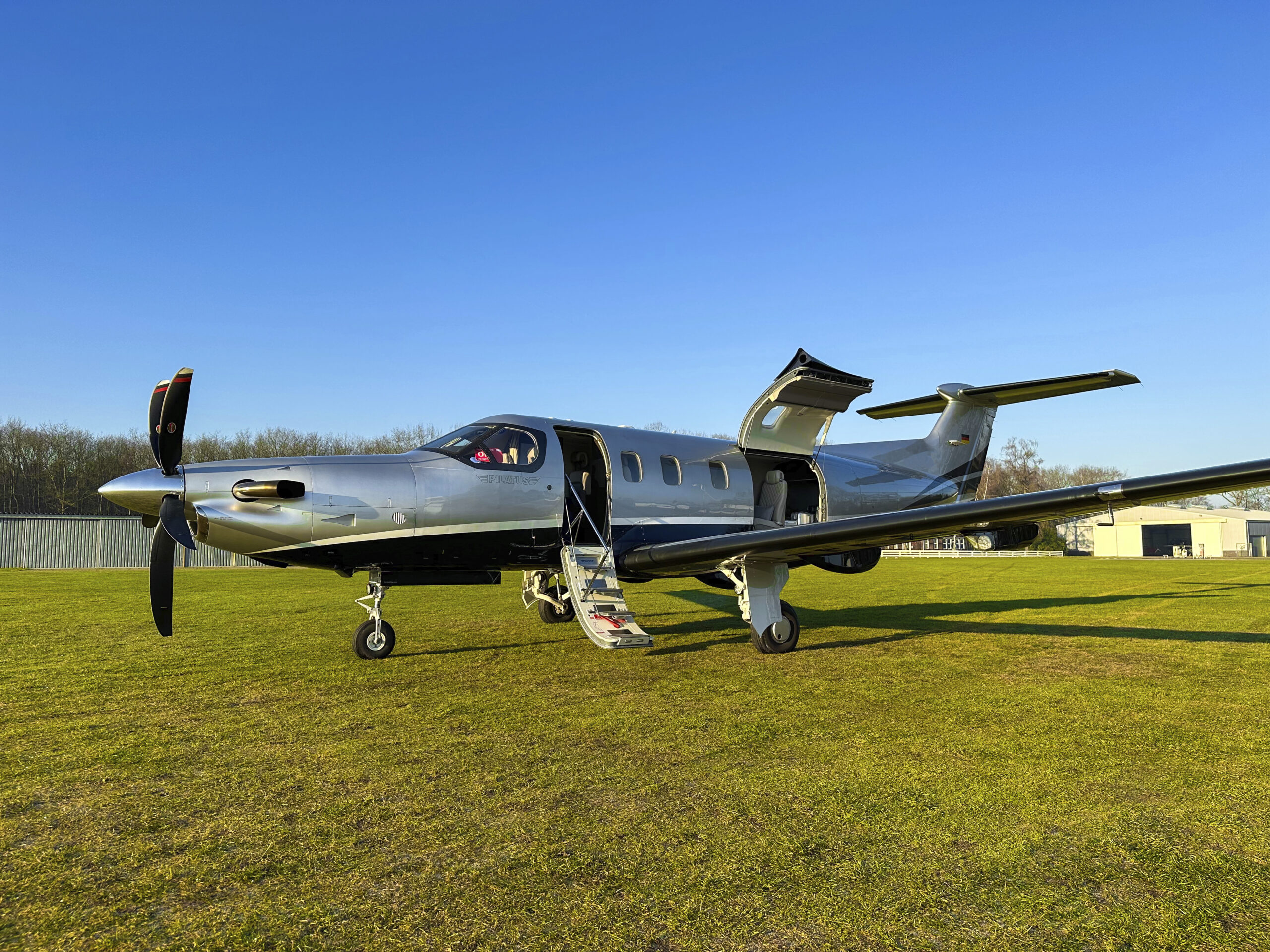 A Lohn Aviation Group Pilatus PC-12 NGX on a grass runway with its cabin door and cargo door open.